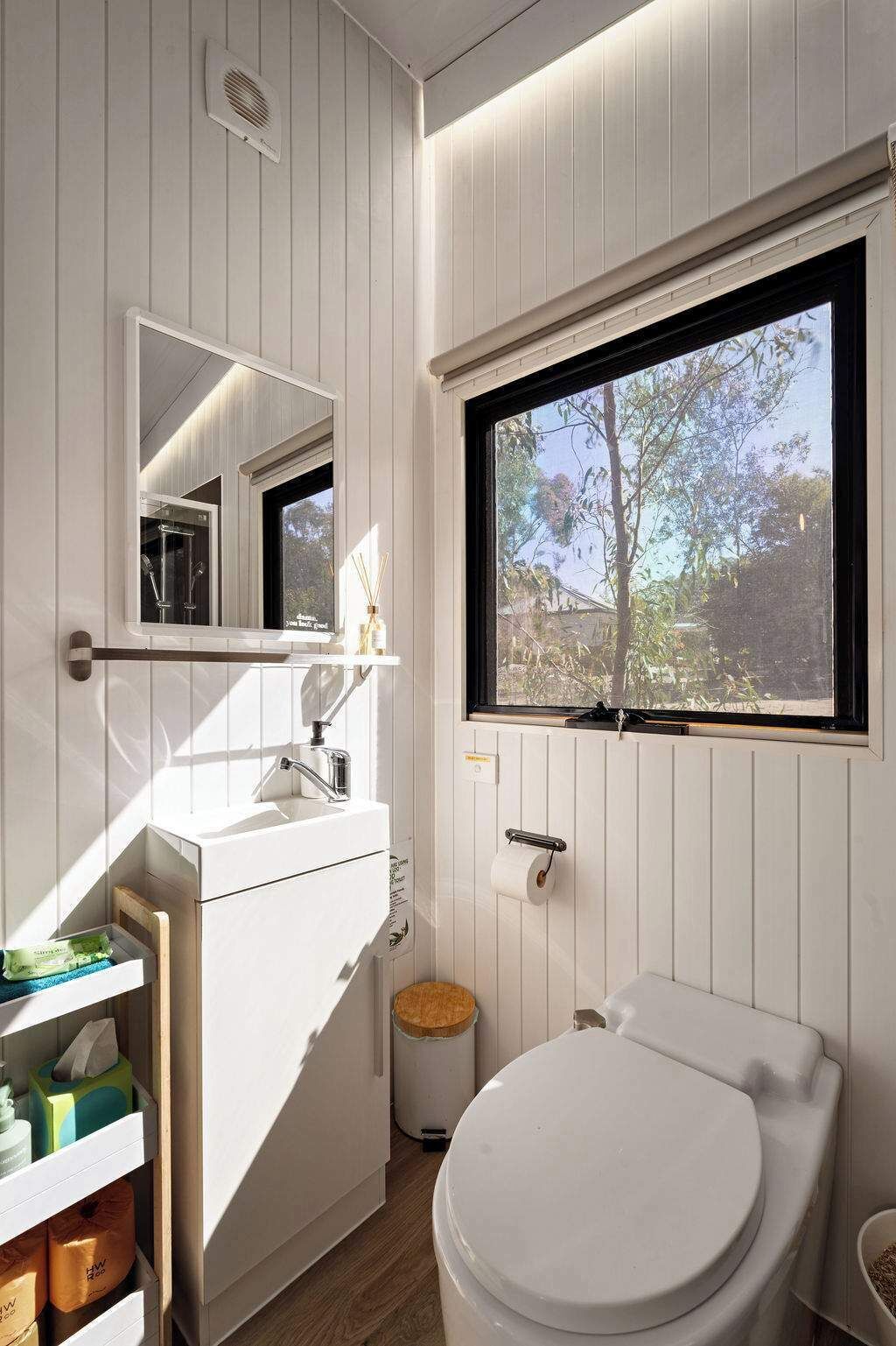 A modern bathroom with a toilet, sink, and mirror. There is a window providing natural light and a view of trees outside.