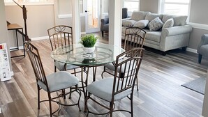A dining area with a glass table surrounded by metal chairs, a wooden floor, and a view of a living room with a sofa and a window.