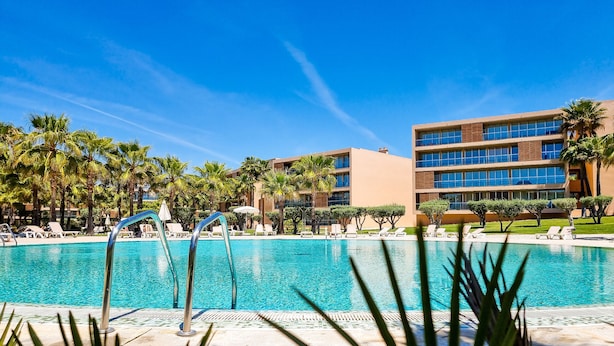 A swimming pool with lounge chairs and palm trees, surrounded by a modern building with multiple floors and large windows. - FLH Salgados Family Apartment (Albufeira)