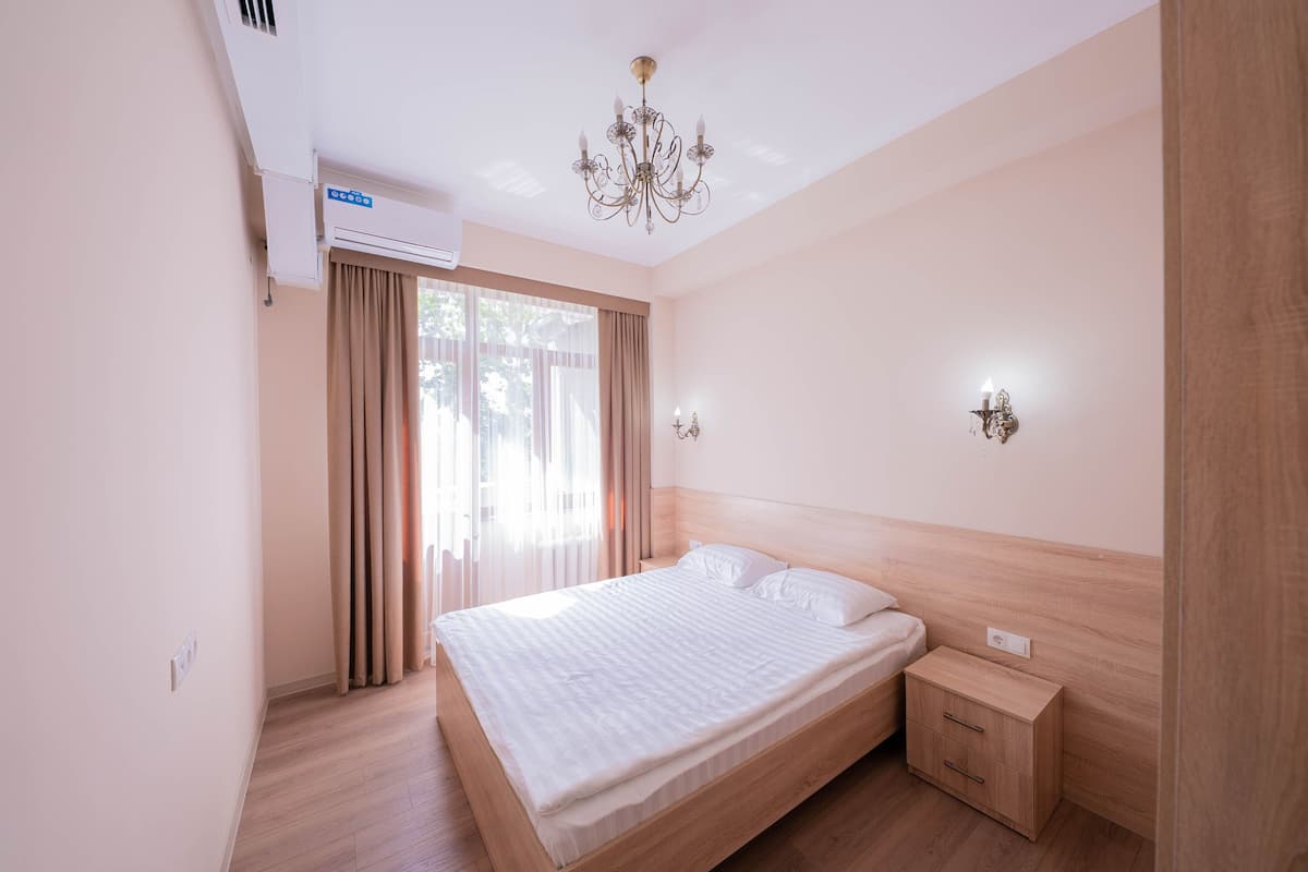 A bedroom with a large bed, a bedside table, a nightstand, a chandelier, and a window with curtains.