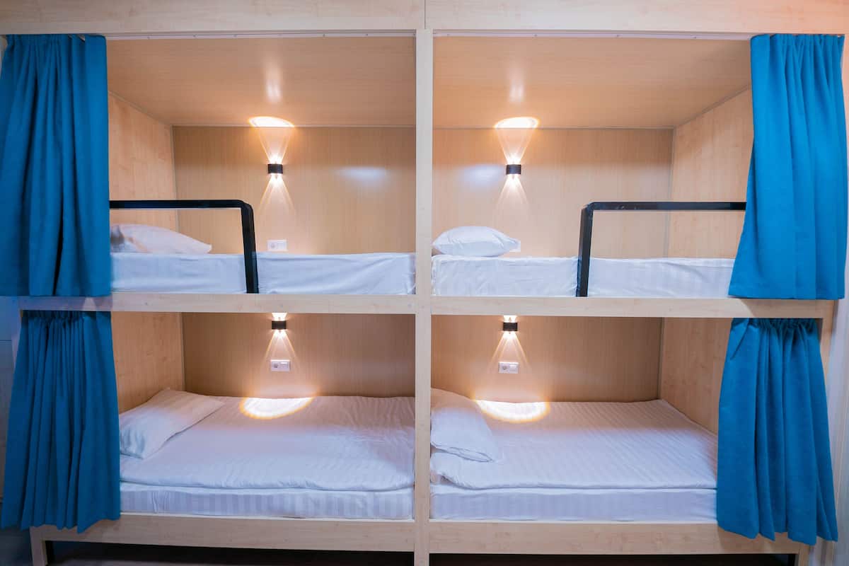 A four-bed wooden cabin with blue curtains, each bed with a white mattress and pillow, and a wall-mounted light fixture.