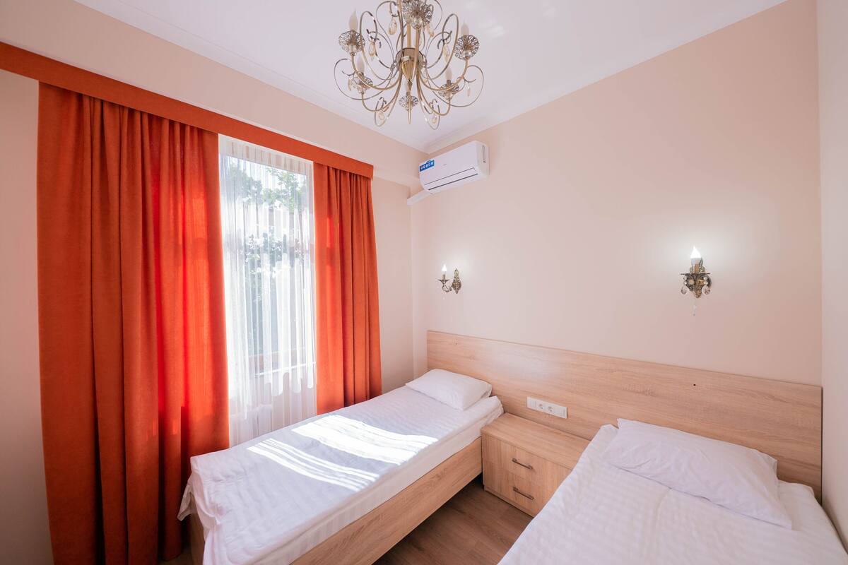A hotel room with two single beds, a wooden headboard, a chandelier, an air conditioning unit, and a window with orange curtains.