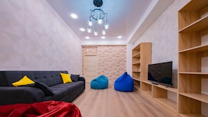 A modern living room with a grey sofa, blue and red bean bags, a TV on a wooden shelf, and a brick wall. - Hostel Kaganat (Bishkek)