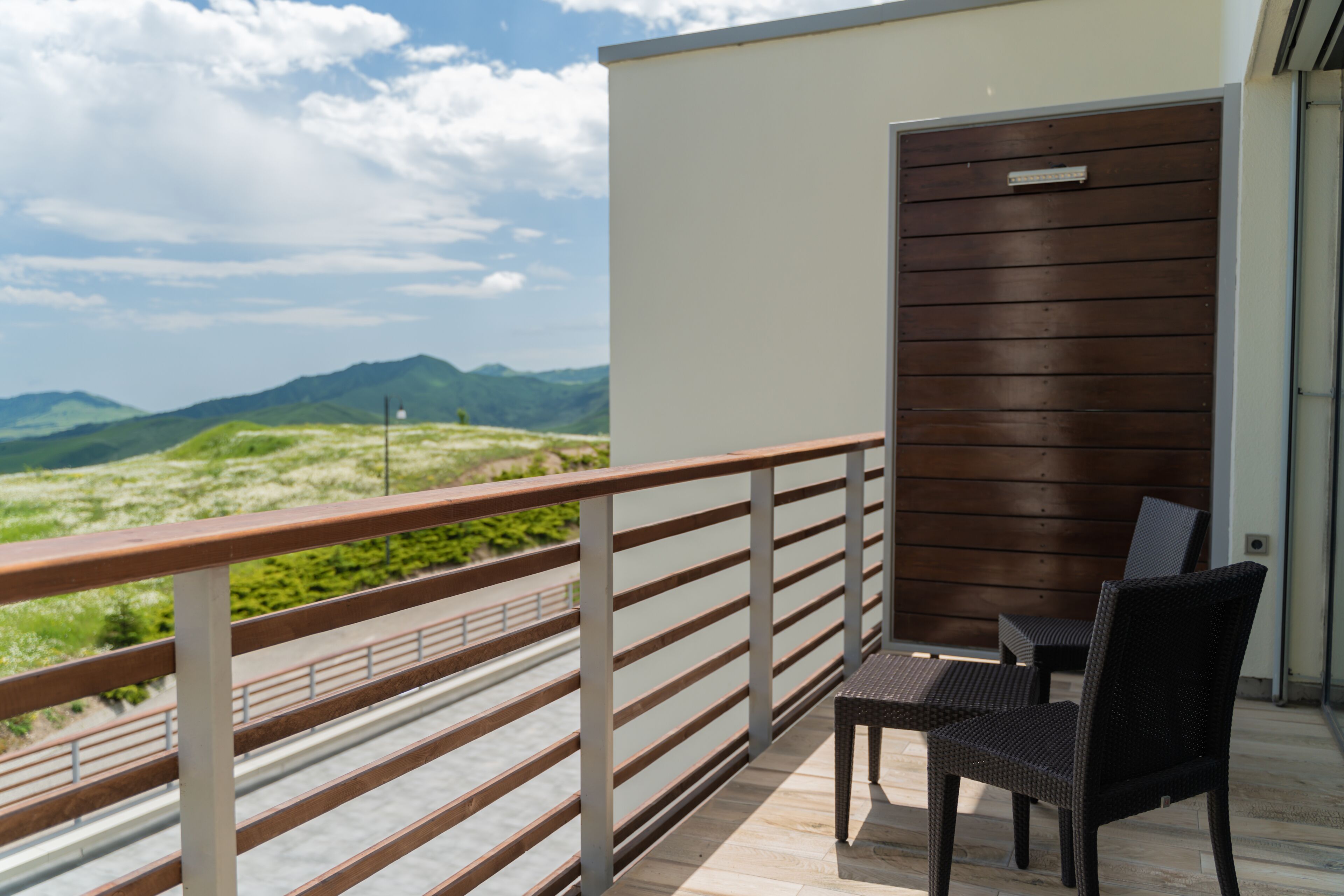 A balcony with a view of green hills, a wooden railing, and two wicker chairs.