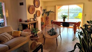 Un salon chaleureux avec un canapé, une table basse et un fauteuil en osier. On y trouve une cheminée ornée d’assiettes décoratives au mur, ainsi qu’un coin repas avec une table et des chaises.
