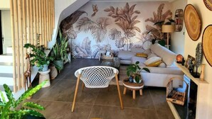 Un salon avec un plafond en pente, un canapé, un fauteuil blanc et une fresque murale représentant des feuilles tropicales.