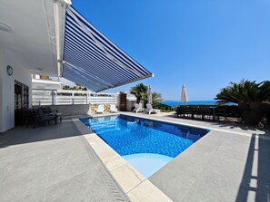Una piscina exterior moderna con vista despejada al mar, rodeada de sillas de descanso y una zona de estar cubierta.