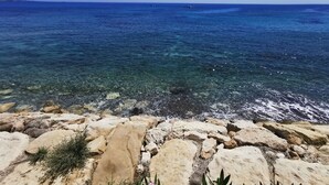 Paisagem costeira com mar azul cristalino, costa rochosa e vegetação rasteira.