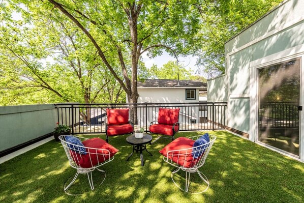 A patio with two red cushioned chairs and a small table, surrounded by a metal fence and lush green grass. - Cozy Boho Guesthouse + Rooftop Deck in the Trees (Austin)