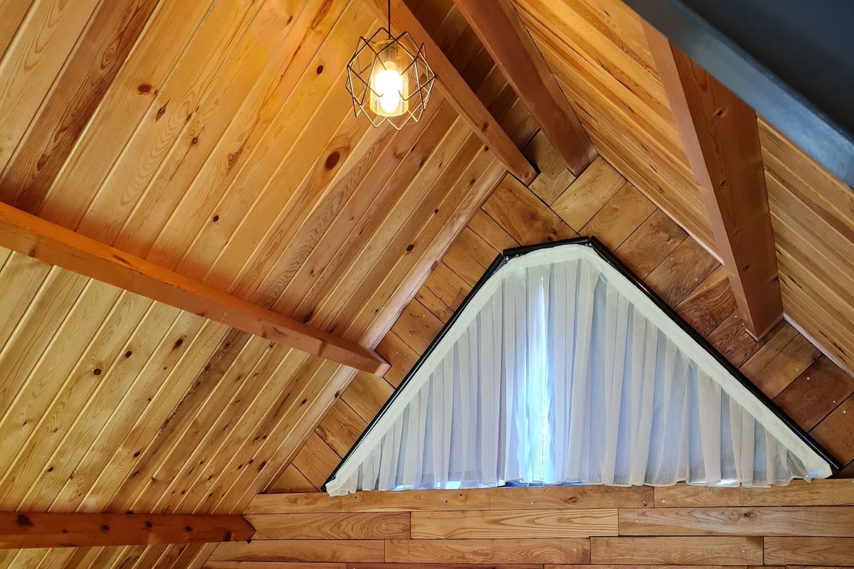 Ett mysigt sovrum på vinden med ett sluttande träpanelat tak, en stor säng med vita sängkläder och en hängande lampa.
