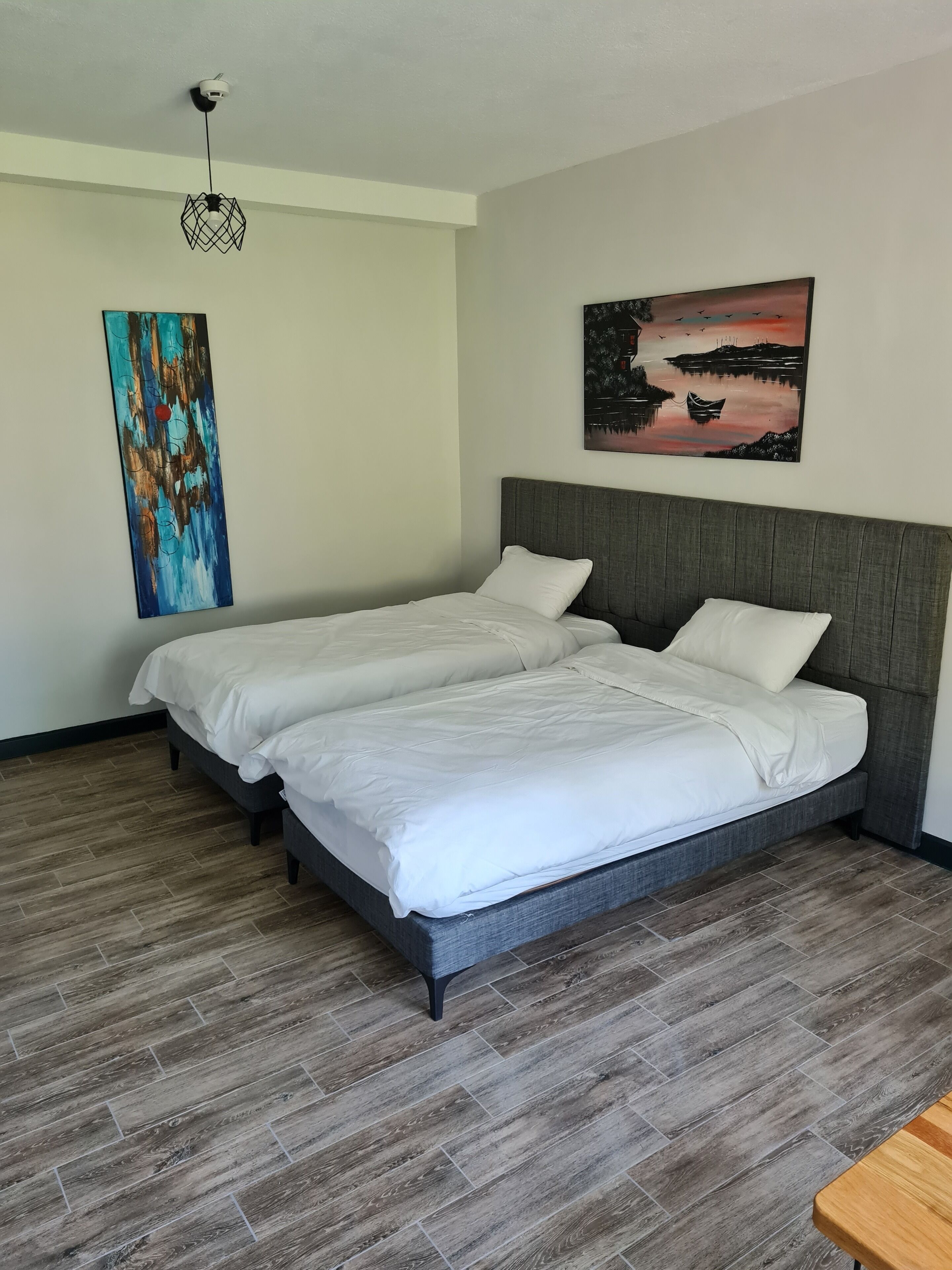 Uma cama de casal com cabeceira cinzenta em quarto de hotel com piso de madeira e uma grande pintura abstrata na parede.