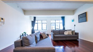 A spacious living room with a large gray sectional sofa, a smaller gray sofa, and a coffee table. The room has hardwood flooring, large windows with curtains, and a ceiling with exposed beams. - Wiltshire | Large & Spacious (Melbourne)