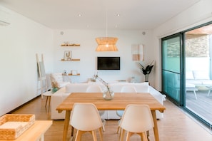 A modern living room with a wooden dining table, white chairs, a sofa, and a TV mounted on the wall.