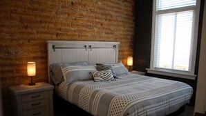 A bedroom with a brick wall, a bed with a white headboard, two bedside tables with lamps, and a window with blinds. - The Lawft, Suite 1,  Essentially 4 bedrooms each with attached Bathroom,No pets! (Russell)