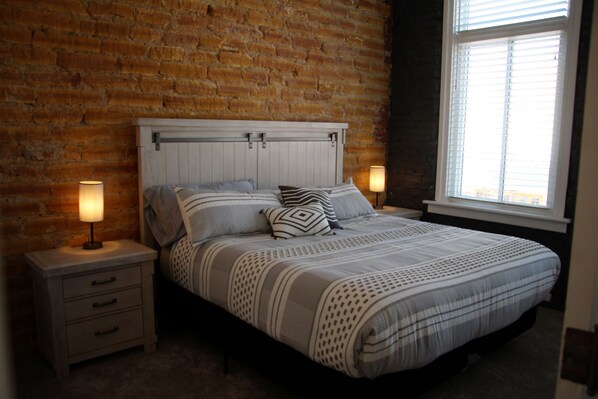 Un dormitorio con pared de ladrillo, una cama con cabecera blanca, dos mesitas de noche con lámparas y una ventana con persianas.