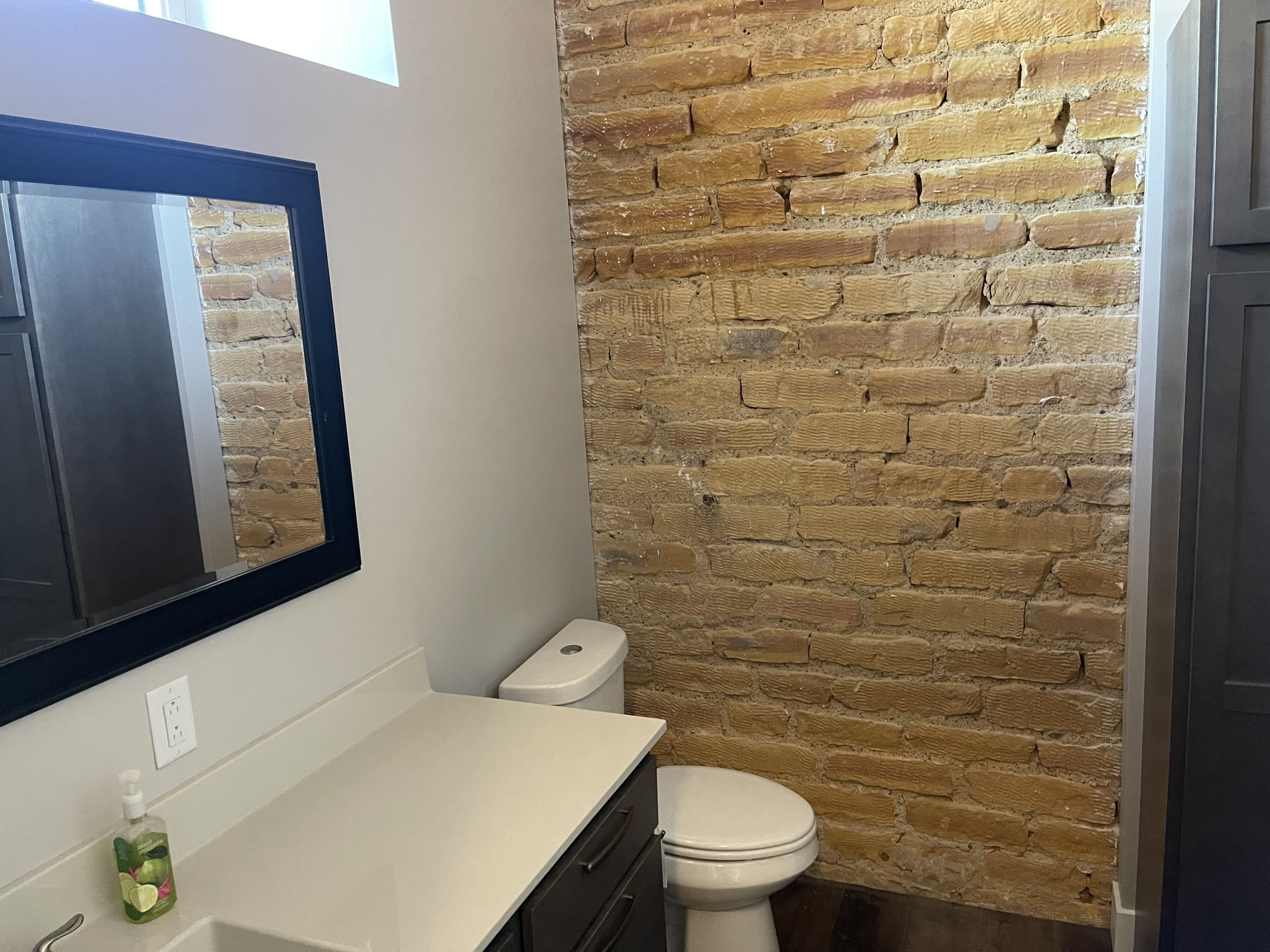 A bathroom with a stone wall, a white toilet, a white sink with a mirror above it, and a bottle of hand soap on the counter.