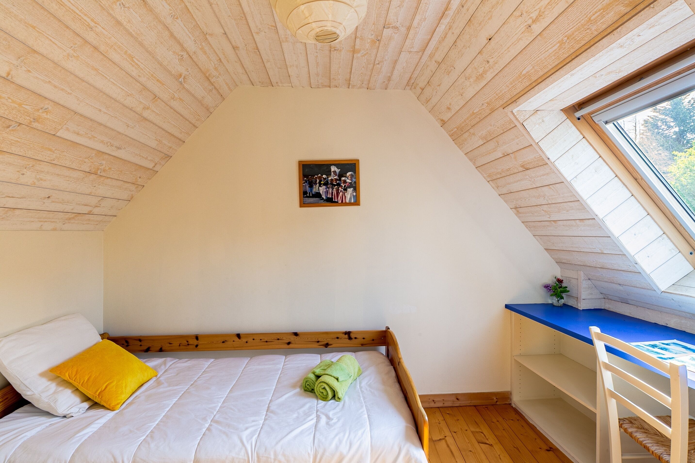 A bedroom with a wooden ceiling, a bed with white bedding and a yellow pillow, a small picture on the wall, and a window with a view of trees.