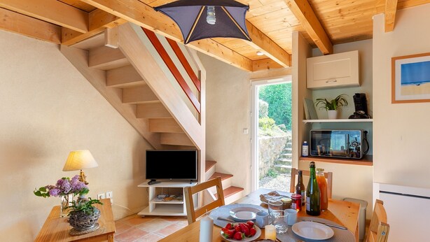A cozy kitchen with a wooden staircase, a dining table set with food, and a TV on a shelf. - Ty ar Hezeg - Gîte 3e avec terrasse privative (Telgruc-sur-Mer)