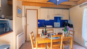 A compact kitchen with wooden cabinets, a blue-tiled backsplash, and a dining area with a wooden table and chairs. - Ty ar Hezeg - Gîte 3e avec terrasse privative (Telgruc-sur-Mer)