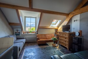 Un appartement-loft chaleureux avec un plafond en pente, une grande fenêtre offrant une vue sur les montagnes, un canapé en cuir marron, une table basse en verre et une commode en bois sur laquelle est posée une télévision.