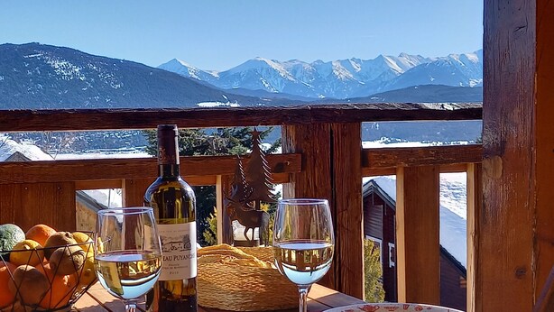 Ein Tisch mit einer Weinflasche, zwei Weingläsern, einem Teller und einem Obstkorb. Ein Holengeländer blickt auf ein verschneites Gebirge.