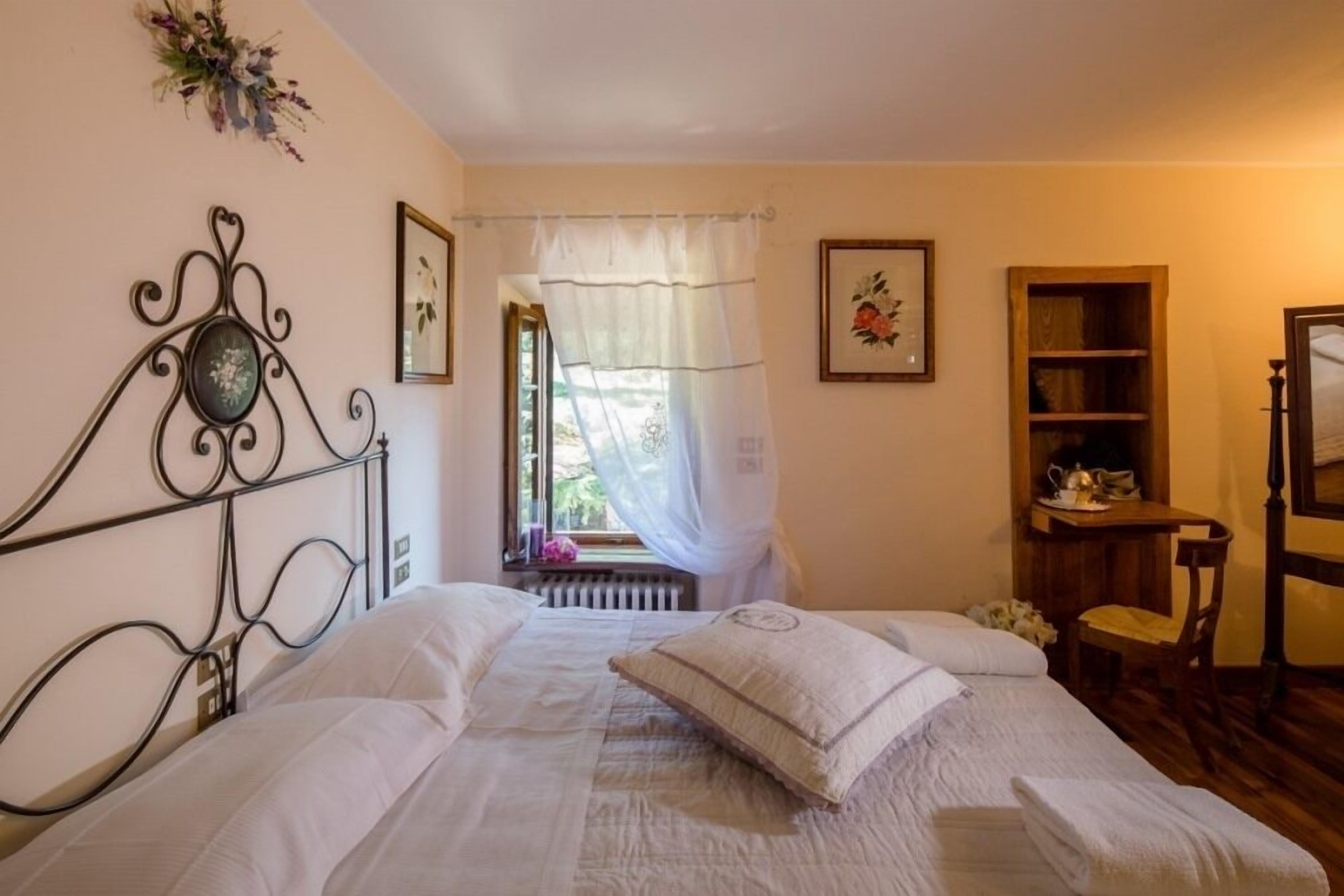 Ein Schlafzimmer mit Bett, Fenster mit Vorhang, Nachttisch mit Lampe und einem Holzstuhl.