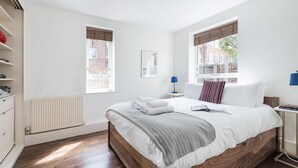 Ein ordentlich eingerichtetes Schlafzimmer mit einem hölzernen Bett, weißer Bettwäsche, einem Heizkörper und zwei Fenstern mit Jalousien.