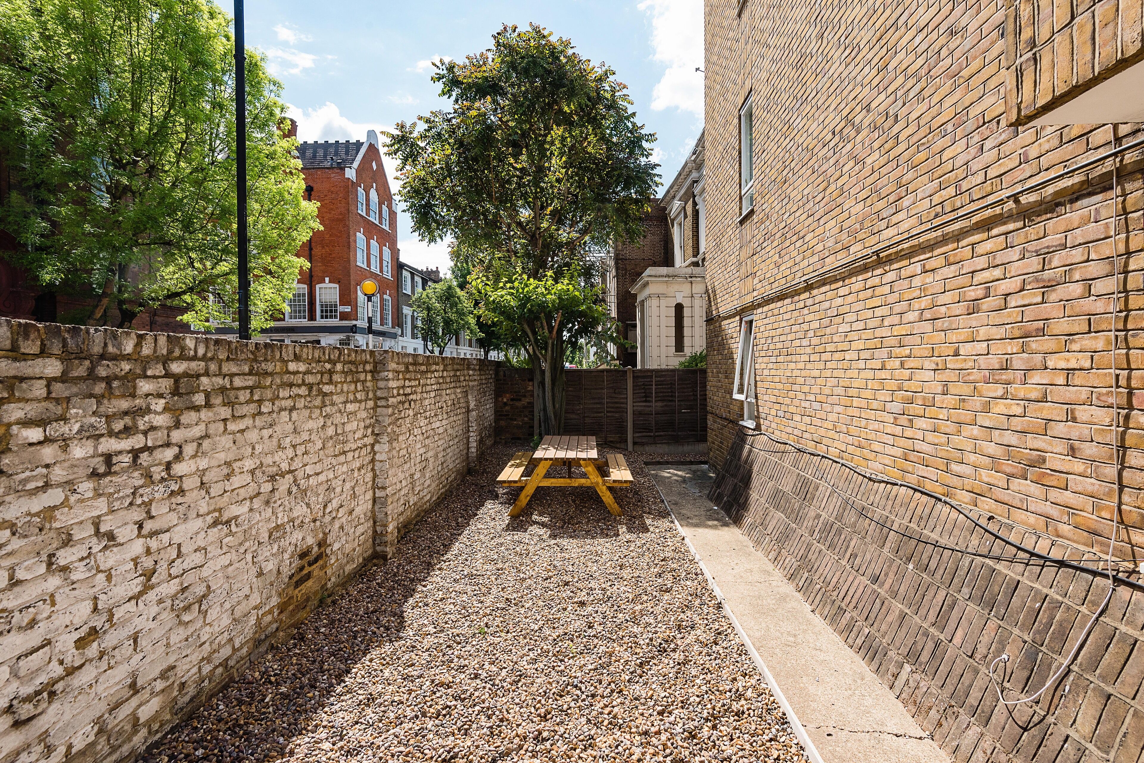 A narrow alley with a gravel path, a wooden picnic table, and a brick wall.