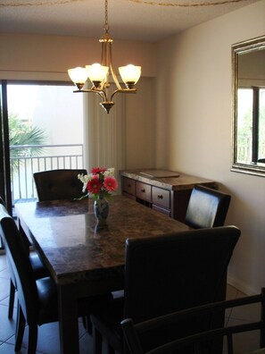 A dining area with a long table, chairs, a vase with flowers, a chandelier, and a window with a view of greenery. - Midnight Cove #532, Bayside in Siesta Key, FL, 2BR-1 1/2 BA, 3rd Floor Walk-Up (Sarasota)