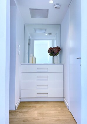 Un couloir étroit avec une commode blanche, un miroir et un vase rempli de fleurs.