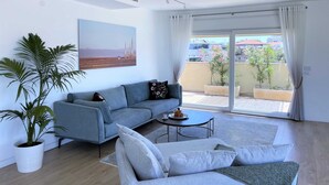 A modern living room with a grey sofa, a white armchair, a coffee table, and a large window with a view of a balcony and buildings. - Seafront penthouse - terrace, AC, lift & parking (Tel Aviv-Yafo)