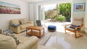A spacious living room with a beige sofa, a wooden coffee table, and a blue ottoman. There is a patterned armchair and a large window providing a view of the garden. - Wheatlands (Hayling Island)
