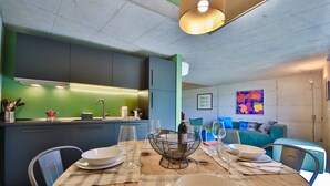A dining area with a wooden table set for four, a kitchen in the background, and a green wall with a colorful painting. - COTTAGE Elena by KlabHouse-4pax w/AC & Lakeview  (Dolo)