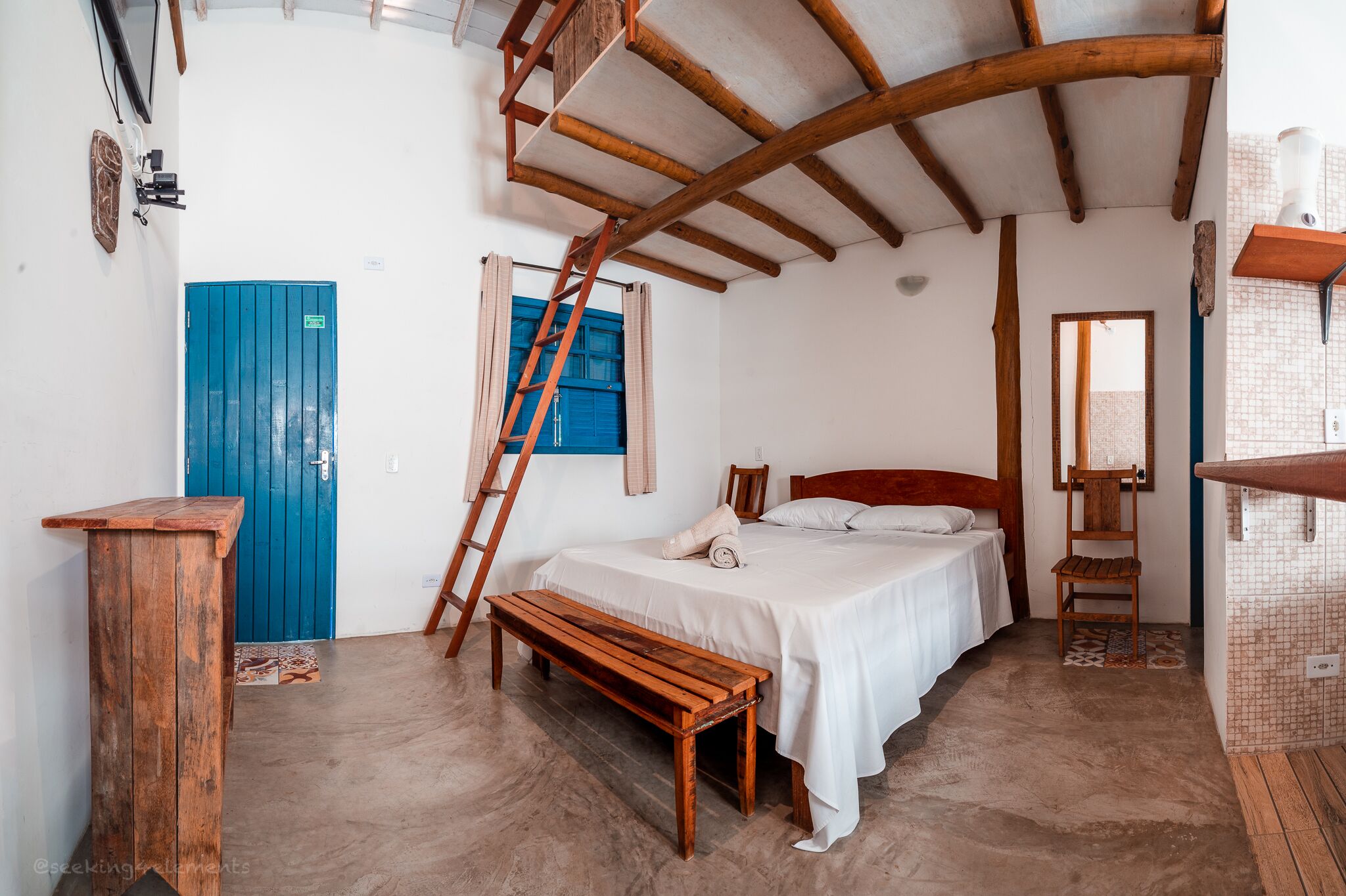 A bedroom with a wooden ceiling, a bed, a bench, a blue door, and a ladder.