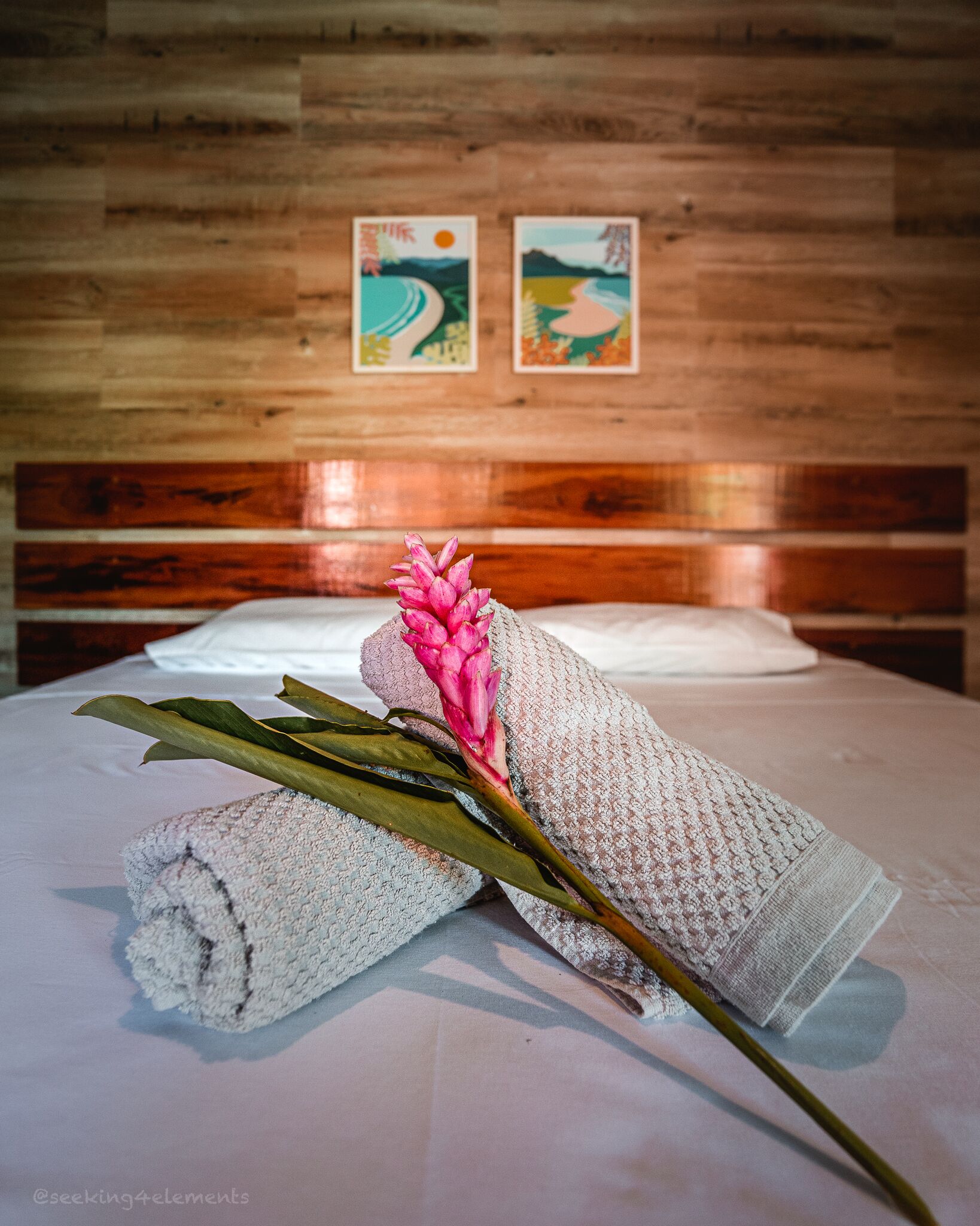 Una cama con una toalla enrollada y una flor rosa encima, frente a una pared de madera con dos cuadros enmarcados.