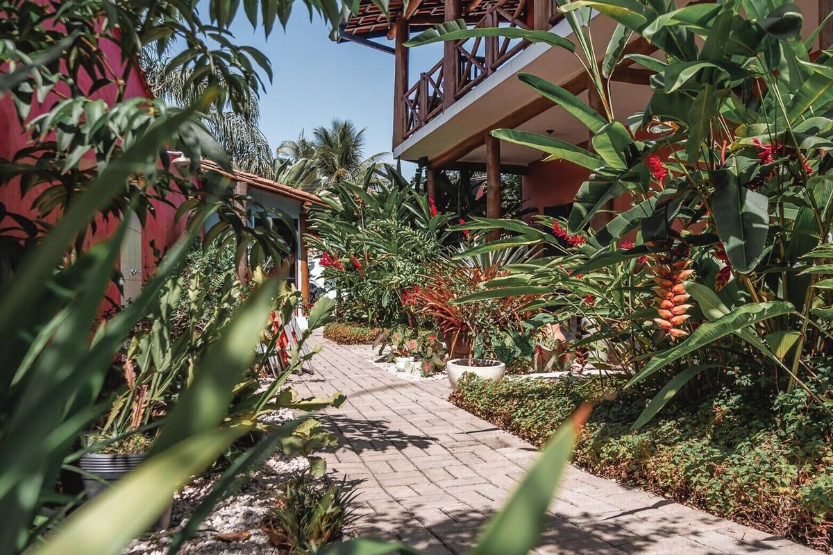 Un sendero conduce a un edificio con balcón de madera, rodeado de exuberante vegetación tropical y un cielo azul despejado.