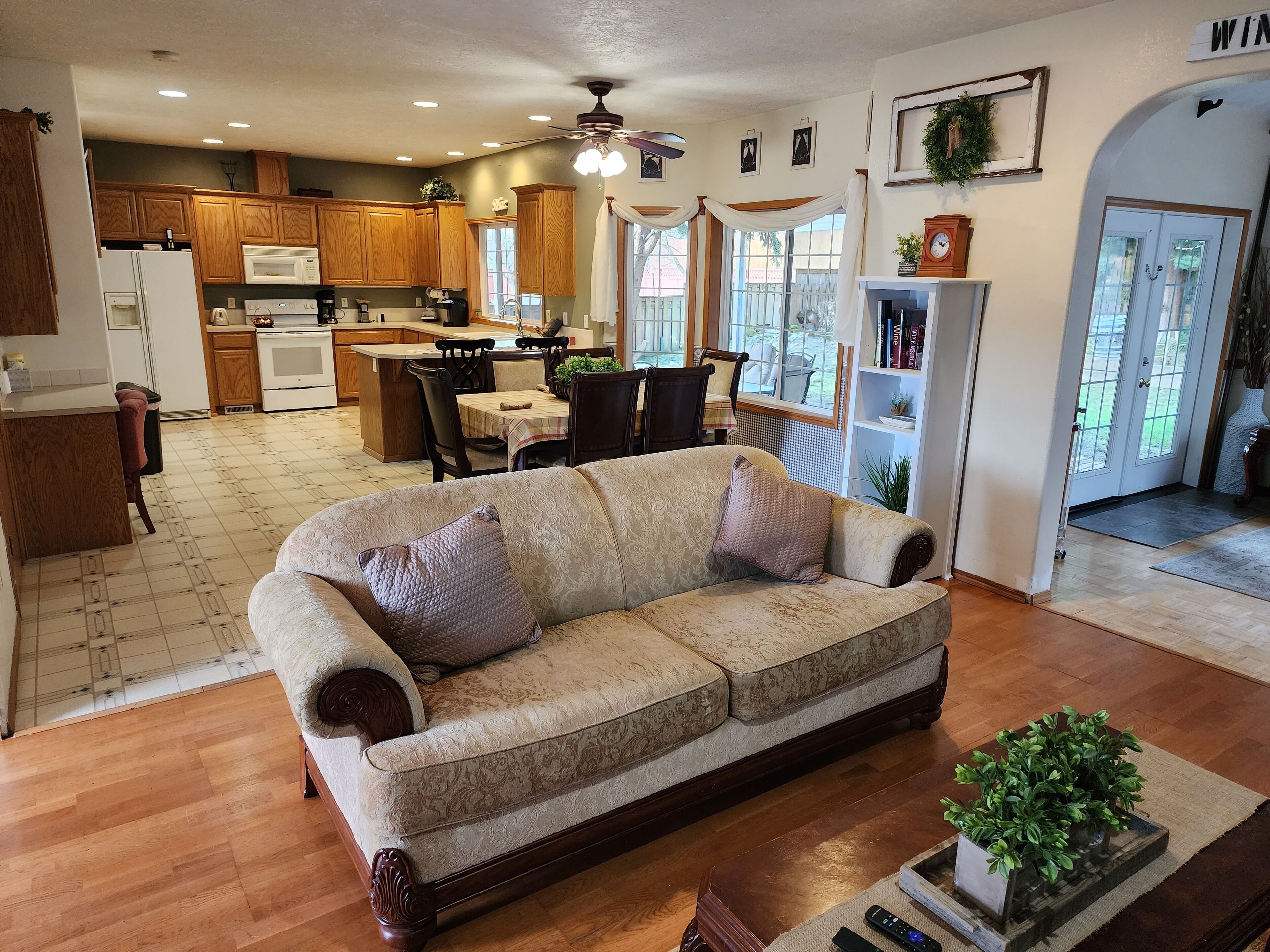 A spacious living room with a large sofa, a dining area with a table and chairs, a kitchen with wooden cabinets, and a ceiling fan.