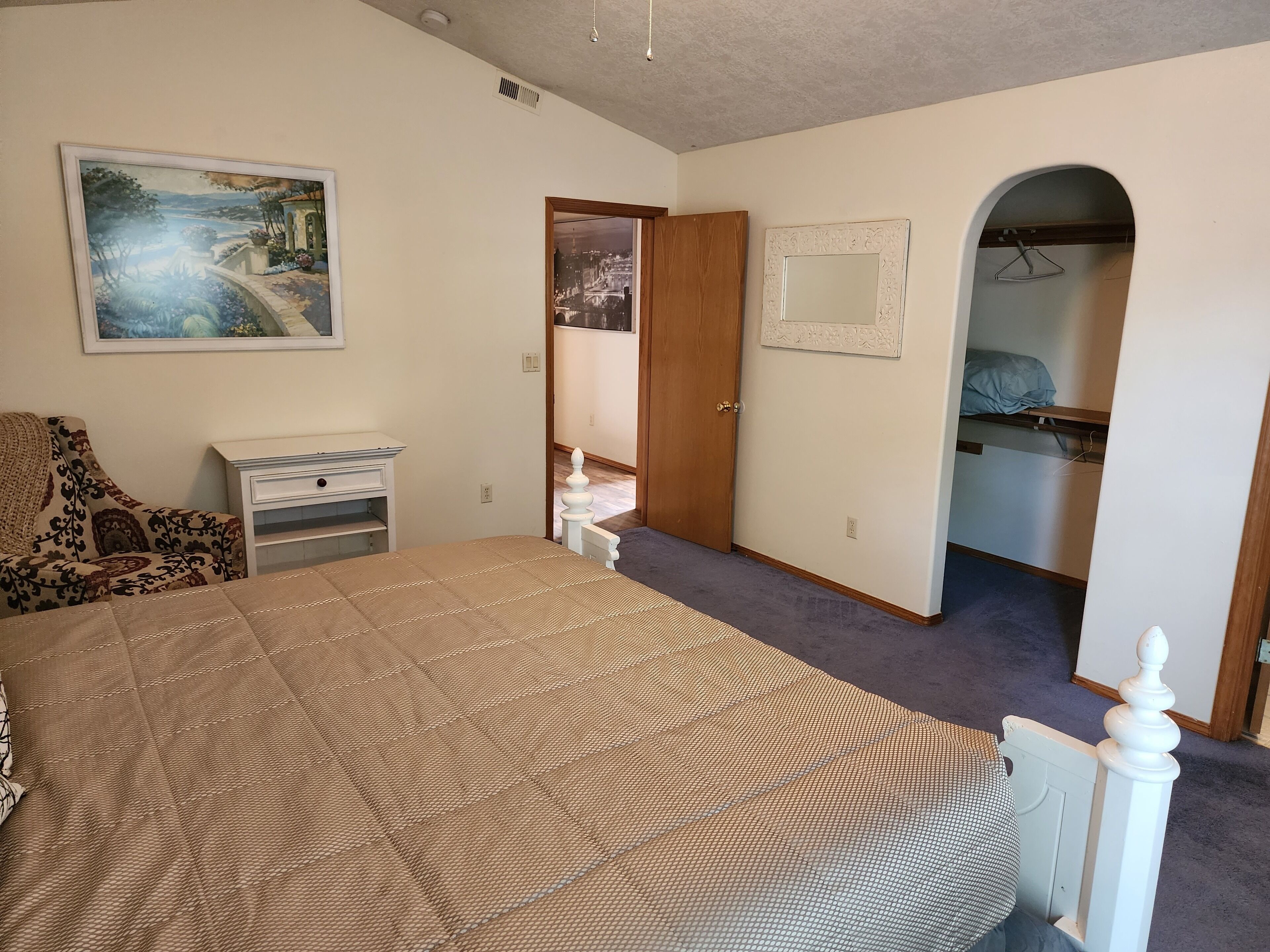 A bedroom with a bed, a dresser, a chair, a painting, a mirror, and an arched doorway leading to a closet.
