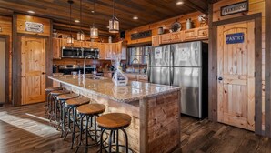 Une cuisine rustique avec des armoires en bois, un îlot central en granit doté de tabourets de bar, des appareils électroménagers en acier inoxydable et des panneaux indiquant « Cuisine du Red Oak Ranch » et « Garde-manger de la ferme ».