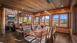 Une salle à manger spacieuse, avec une table et des chaises en bois, une cheminée et de grandes fenêtres offrant une vue sur les montagnes.
