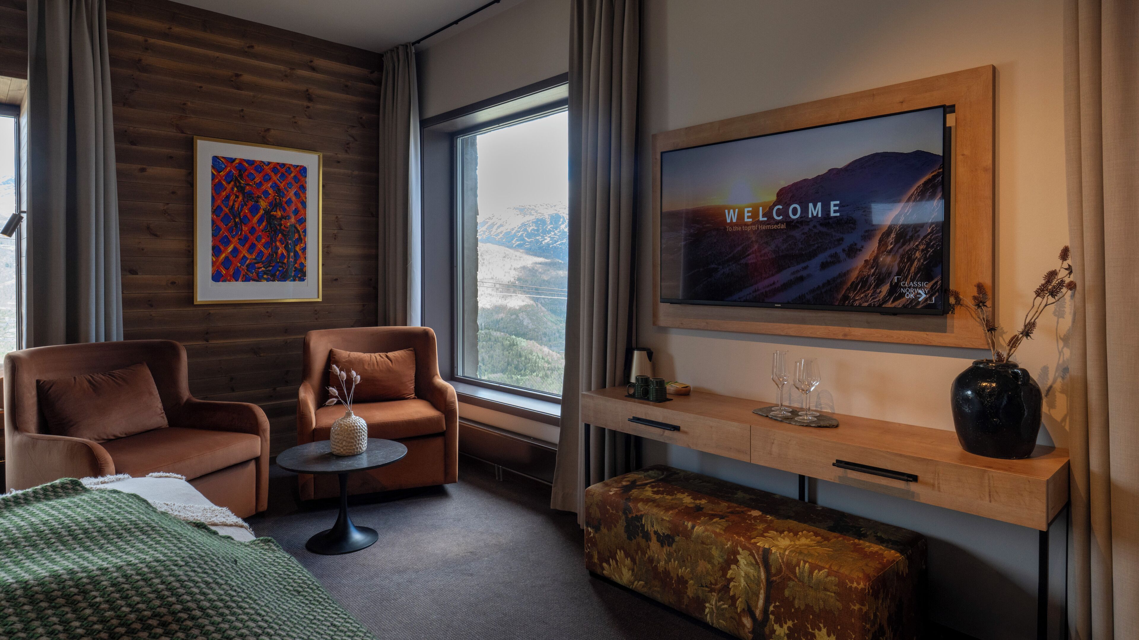 Quarto de hotel moderno com cama, dois sofás, mesa de centro, TV exibindo uma mensagem de boas-vindas e vista para montanhas pela janela.