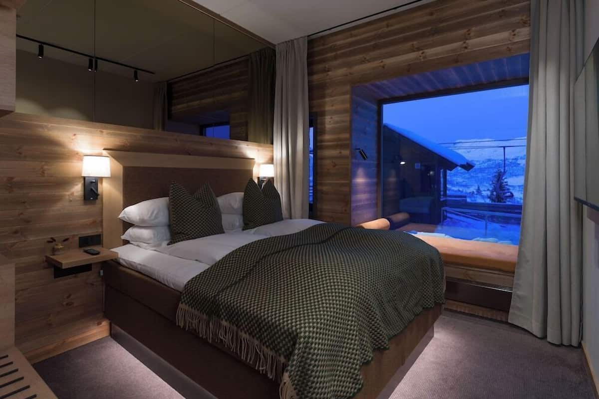 Un dormitorio moderno con cabecera de madera, una cama grande con manta verde estampada, lámparas de noche y vistas a un paisaje nevado a través de un ventanal amplio.