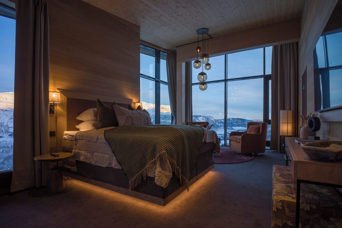 Un dormitorio moderno con una cama grande, un área de descanso y vistas a montañas nevadas a través de amplios ventanales.