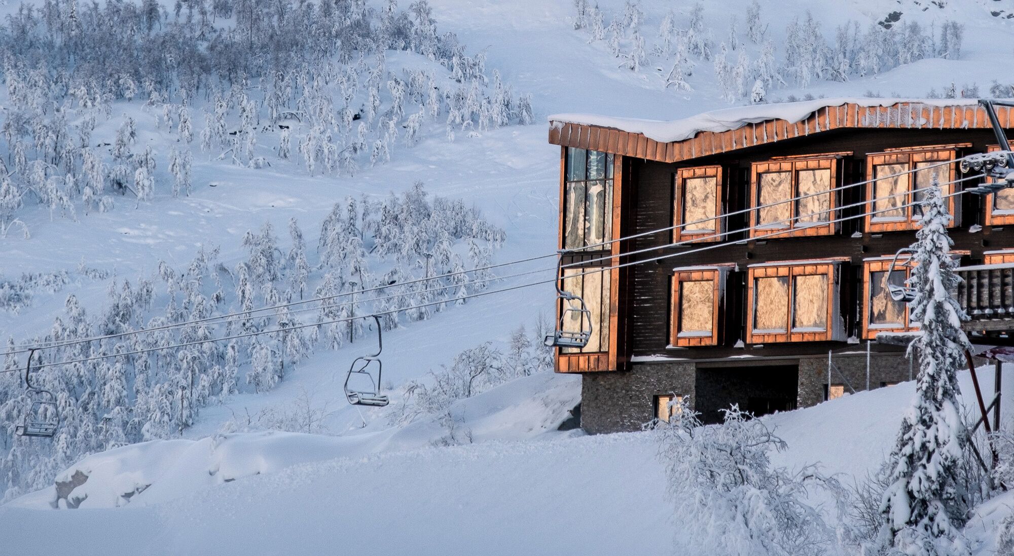 Een houten chalet met meerdere ramen, een skilift op de achtergrond en sneeuwbedekte bomen.