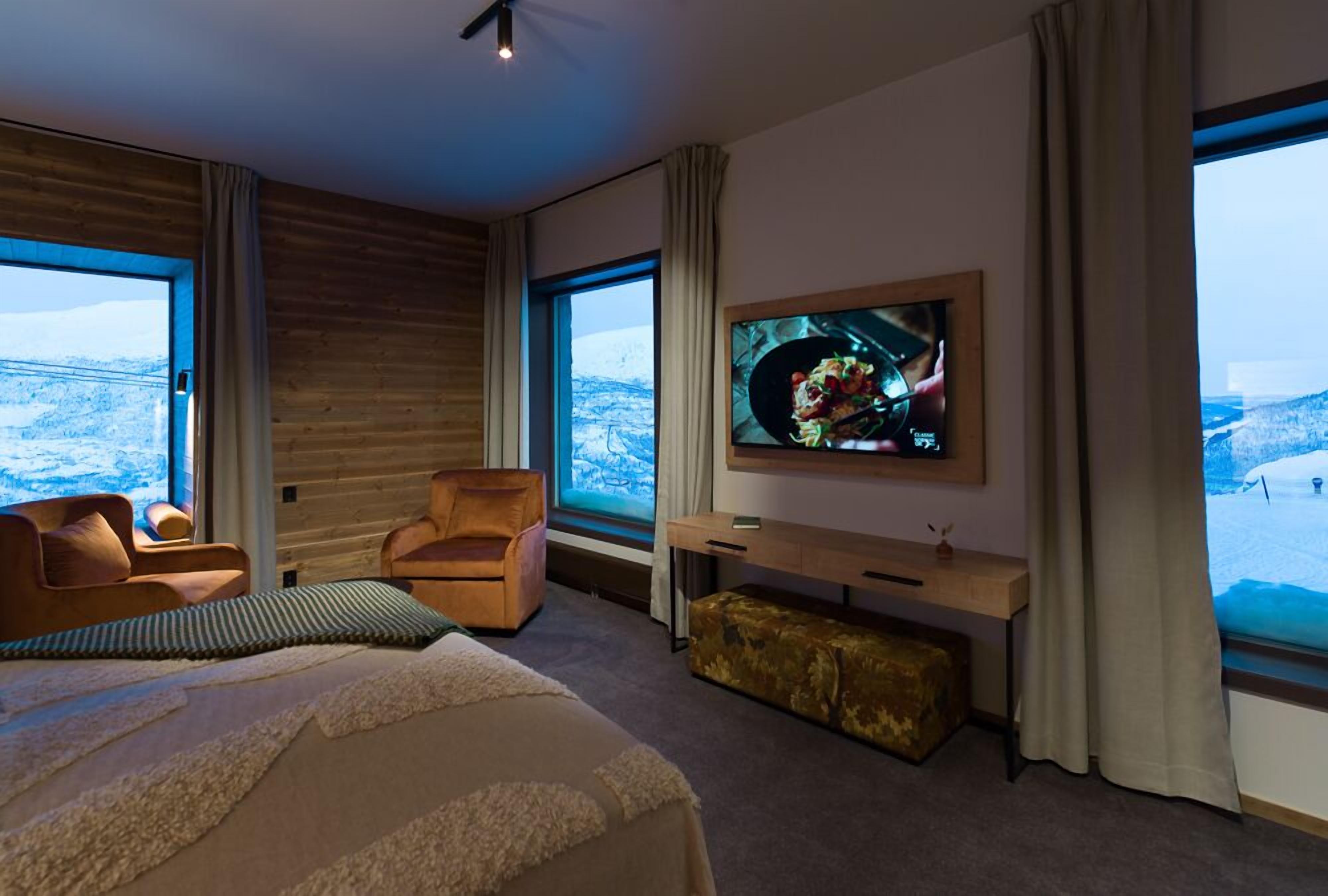 A modern hotel room with a large bed, a TV mounted on the wall, a desk, and a view of a snowy landscape through large windows.
