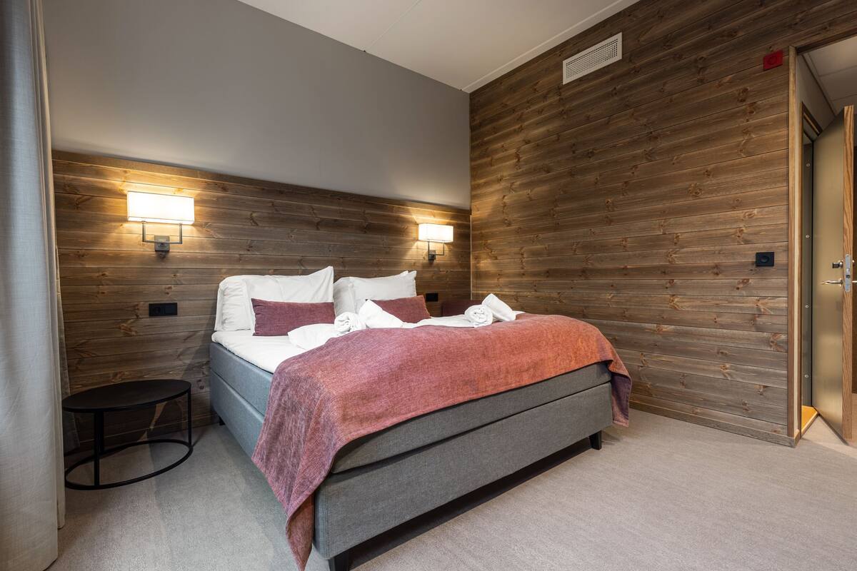 Un dormitorio moderno con una cama, dos luces fijadas a la pared, una mesita redonda y un panel de pared de madera.