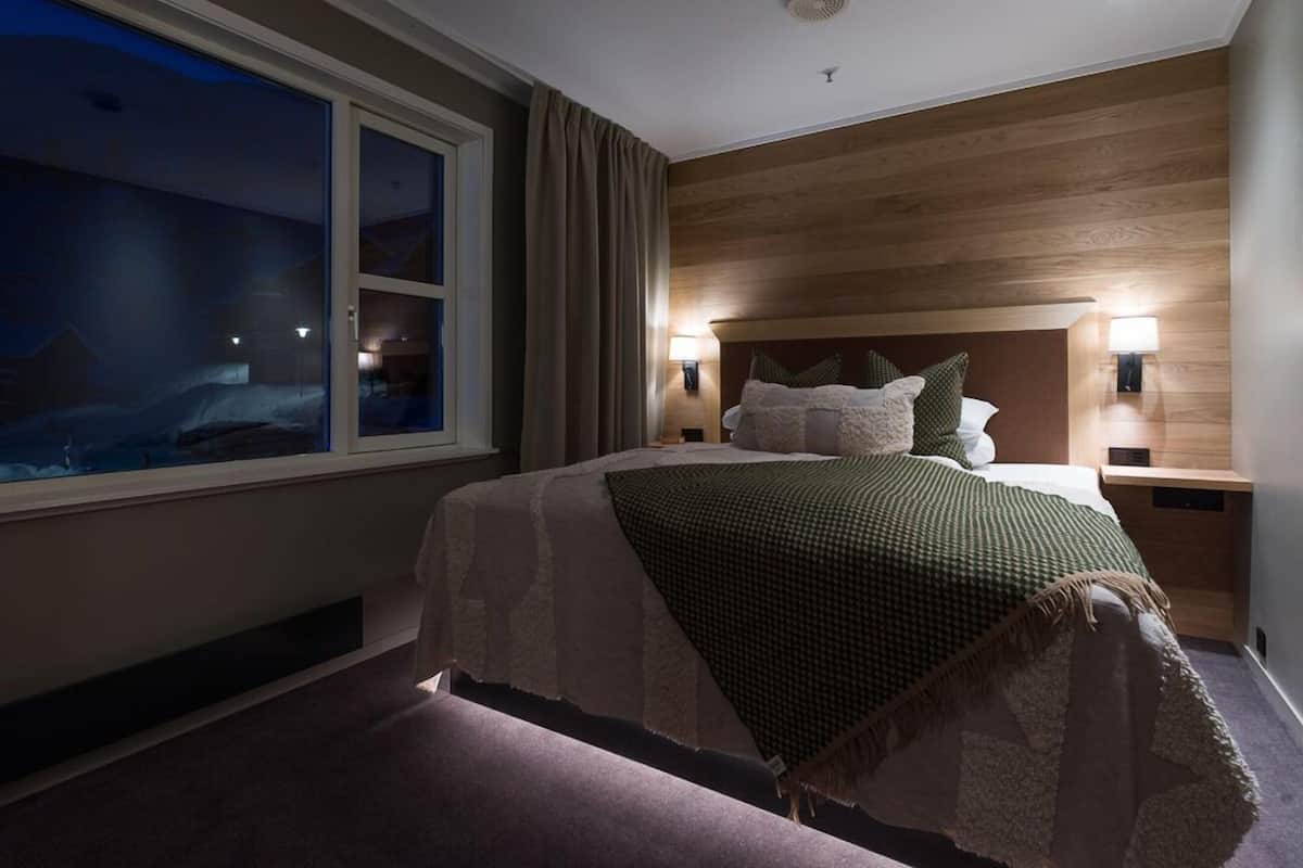 Habitación de hotel con un ventanal grande, una cama con manta verde, paneles de madera en las paredes y dos lámparas de noche.