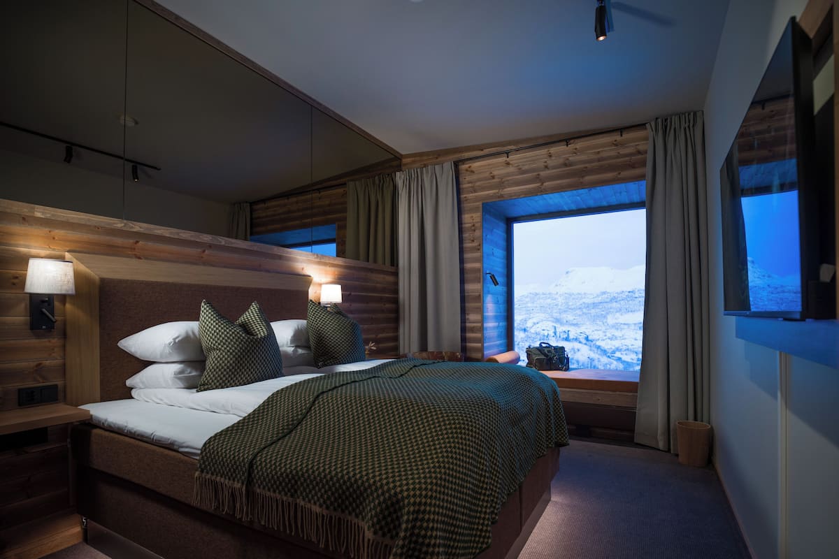 Habitación de hotel moderna con una cama grande, cabecera de madera y vistas a un paisaje nevado a través de un ventanal.