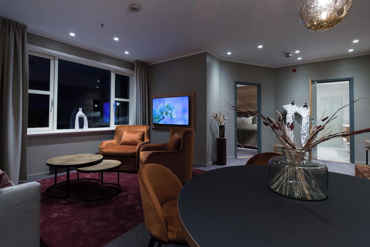 Una sala de estar moderna con televisor, asientos cómodos, una mesa grande y vistas al exterior a través de un ventanal.