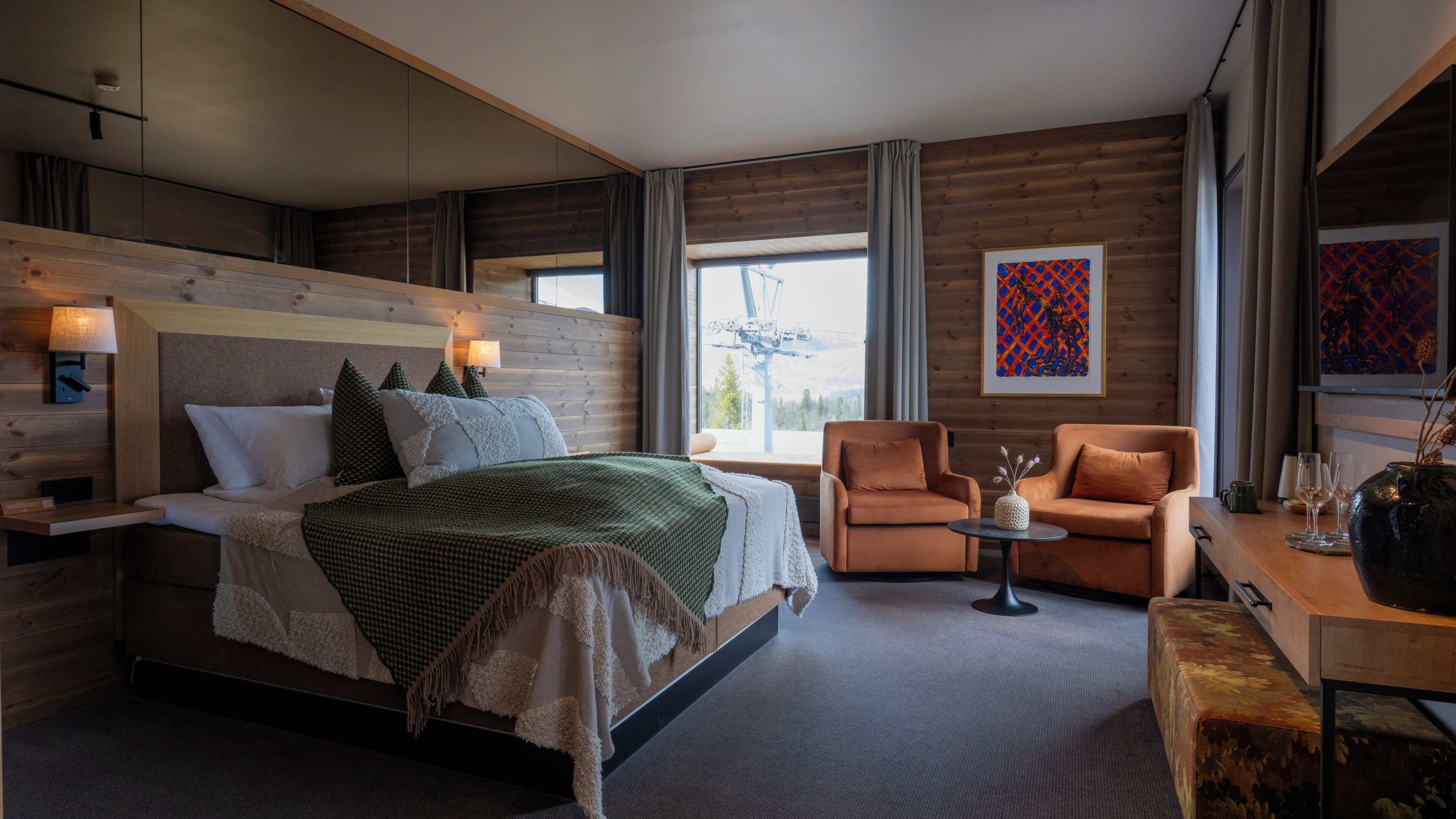 A modern hotel room with a large bed, two armchairs, a small table, and a view of the mountains.